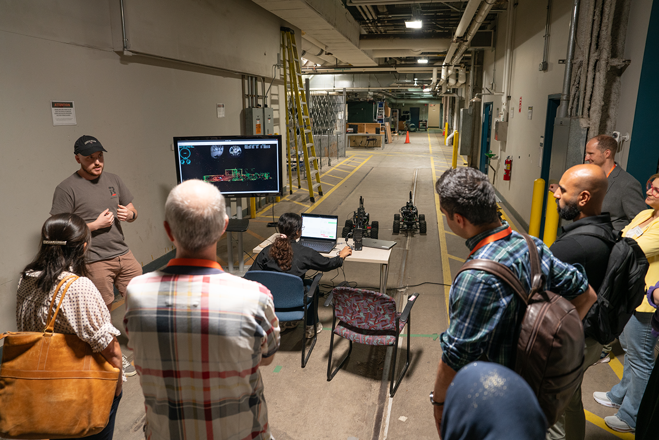 Image of a group of adults watching a demonstration of a two small robotic vehicles on a small indoor track